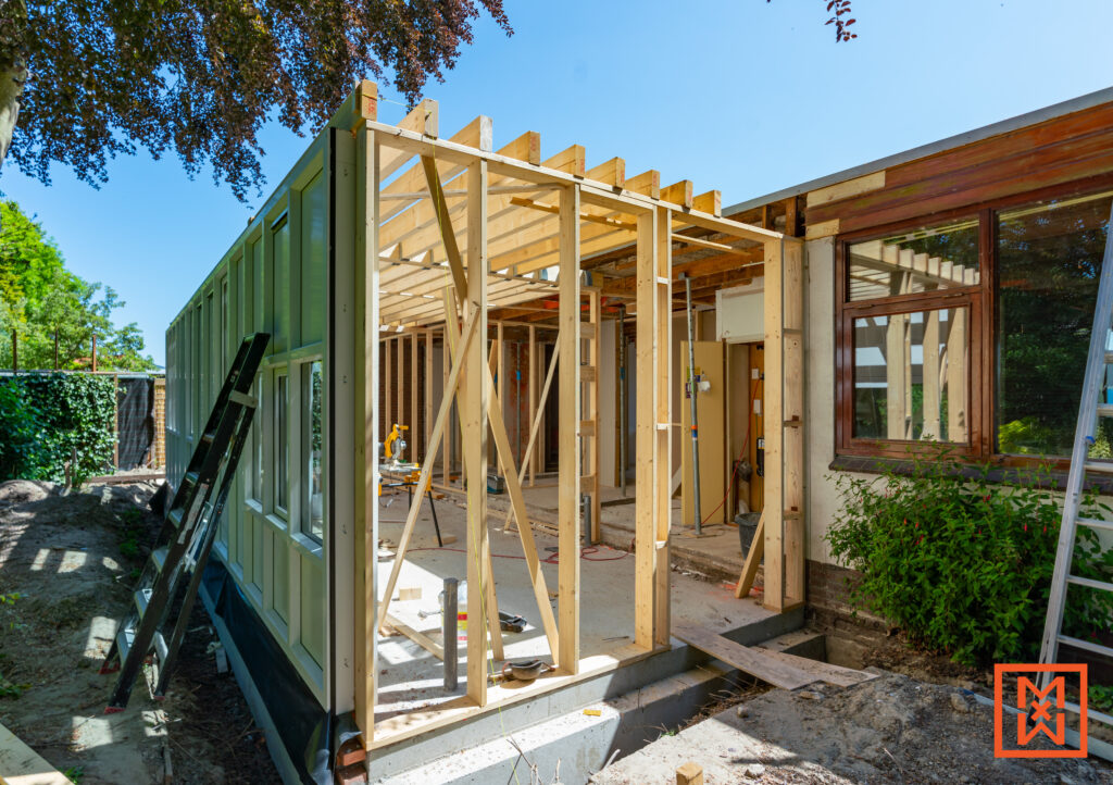 Huis uitbreiding in aanbouw. Houten frame van een aanbouw, met zicht op de bestaande woning en tuin. Bouwen van een nieuwe kamer, met timmerwerk in volle gang.
