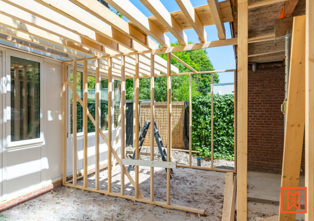 Houten constructie van een veranda in aanbouw. De open houten frame staat tegen een muur van baksteen, met een glazen deur en ramen zichtbaar. Zonnig weer, ideaal voor doe-het-zelf bouwen in de tuin.