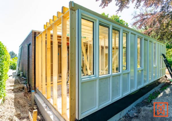 Huis verbouwen in volle gang: houten frame van een nieuwe aanbouw, met ramen en witte gevelbekleding. Een gedeelte van de bestaande woning is te zien, met bakstenen muur en een tuin op de achtergrond.