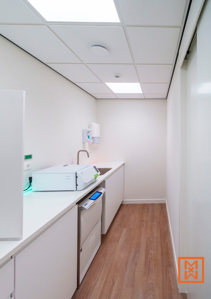 Moderne tandartspraktijk sterilisatieruimte met witte kasten, een wastafel en sterilisatie apparatuur. De vloer is voorzien van houten look, wat een warme uitstraling geeft. Hygiëne in de tandartspraktijk.