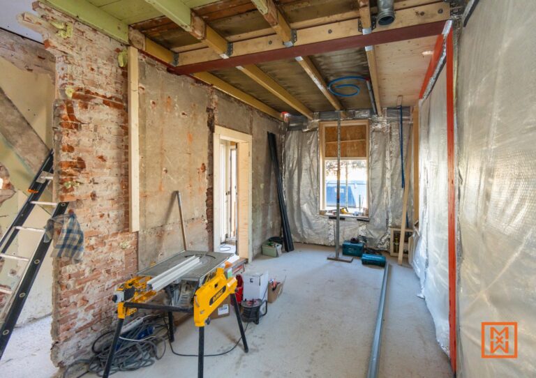 Interieur in verbouwing, met zicht op bakstenen muur, zaagtafel en isolatiemateriaal. De ruimte is in renovatie en er staan gereedschappen klaar om te klussen aan het huis.