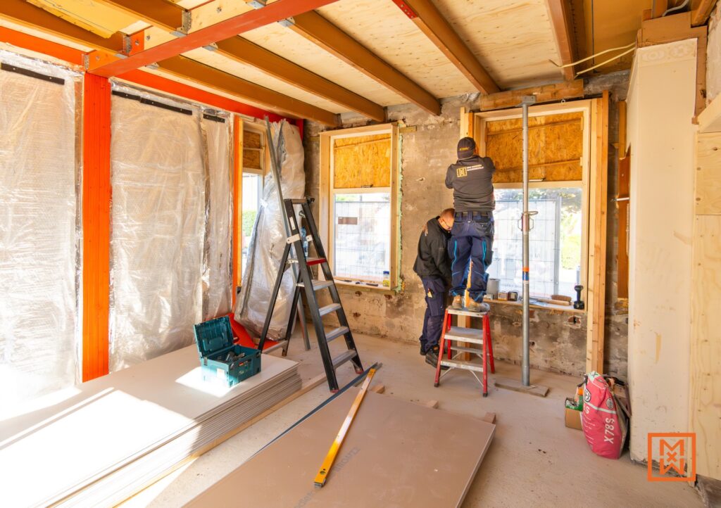Twee bouwvakkers in actie tijdens een renovatieproject. Een man staat op een steiger, de ander op een trapje, bezig met het plaatsen van gipsplaten bij een raam. Zichtbaar zijn ook houten balken, stalen constructie en bouwmateriaal.