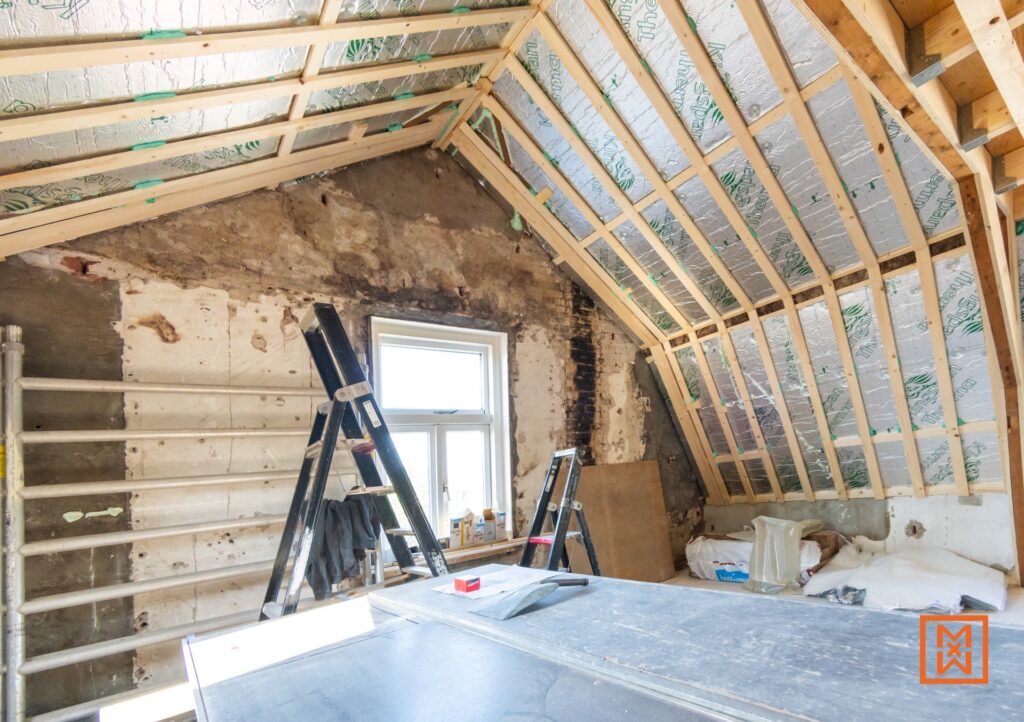 Zolderrenovatie in volle gang: open constructie met houten balken en dakisolatie. Ruwe muren en een ladder wijzen op de beginfase van het klussen. De zolder wordt verbouwd.
