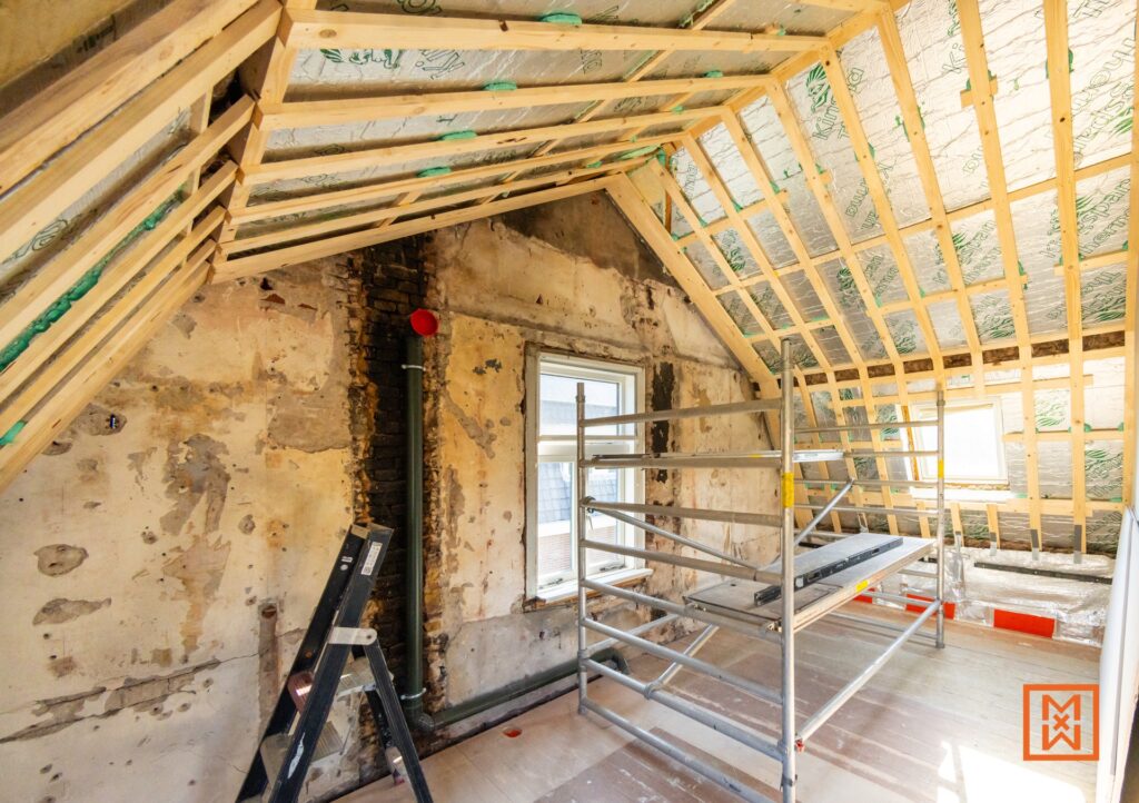 Zolder in aanbouw met houten frame, isolatie en steiger. De oude bakstenen muur is zichtbaar, klaar voor afwerking en isolatie. Project voor zolder verbouwen.