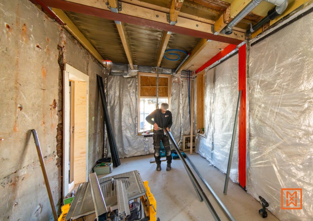 Een man bezig met klussen in een huis dat in renovatie is. Hij snijdt metaal met gereedschap, omringd door isolatiemateriaal. De ruimte is in verbouwing, met zichtbare balken en een zaagtafel.
