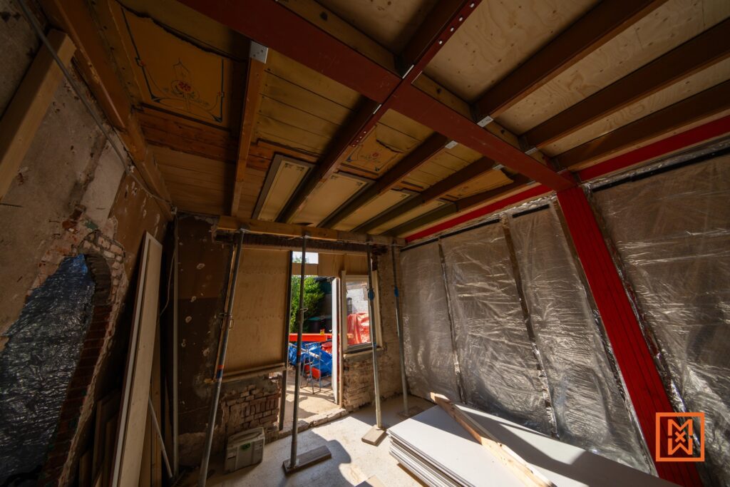 Interieur van een huis in verbouwing. Rode stalen balken ondersteunen de houten constructie van het plafond. Zilveren isolatie en gipsplaten liggen klaar voor renovatie. De bouwwerkzaamheden zijn in volle gang voor een succesvolle verbouwing.