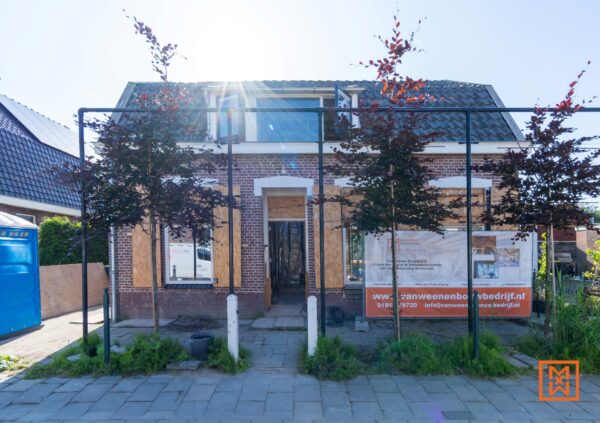 Huis in Nederland in renovatie, met houten platen voor de ramen. Een bouwbedrijf, Van Weenen Bouwbedrijf, heeft een banner op het hekwerk. De zon schijnt fel. Tekst: "Van Weenen Bouwbedrijf" en "huis renovatie".
