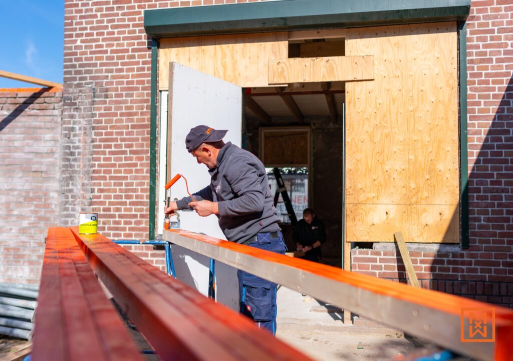 Man schildert een houten balk met oranje verf tijdens een huis renovatie. De klusser draagt een pet en staat voor een bakstenen muur met een open deur. Achter hem is een andere persoon aan het klussen.