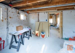 Interieur in verbouwing, met witte muren en een betonnen vloer. Een provisorische werktafel staat klaar, met materialen voor renovatie, zoals hout en gereedschap. De open ruimte suggereert een toekomstig droomhuis.