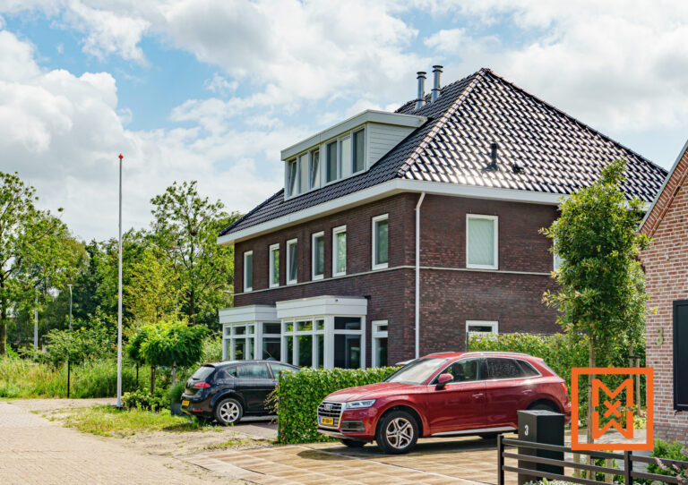 Modern huis van rode baksteen met dakkapellen en zwarte dakpannen, twee auto's geparkeerd op de oprit en een weelderige groene tuin. Op de achtergrond staat een vlaggenmast met een rode top.