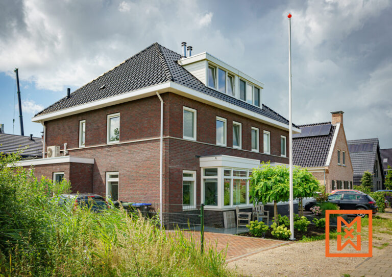 Een modern bakstenen huis met een donker pannendak en dakkapellen staat aan een met bomen omzoomde straat. Het huis beschikt over een serre en een vlaggenmast, met andere huizen zichtbaar op de achtergrond. De tuin bestaat uit een kleine groene boom en een goed onderhouden gazon.