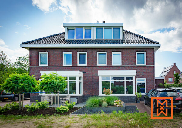 Een prachtig bakstenen huis met een dakkapel en een zorgvuldig aangelegde tuin. Het huis heeft grote witte ramen en een modern design. Er staan twee auto's op de oprit.