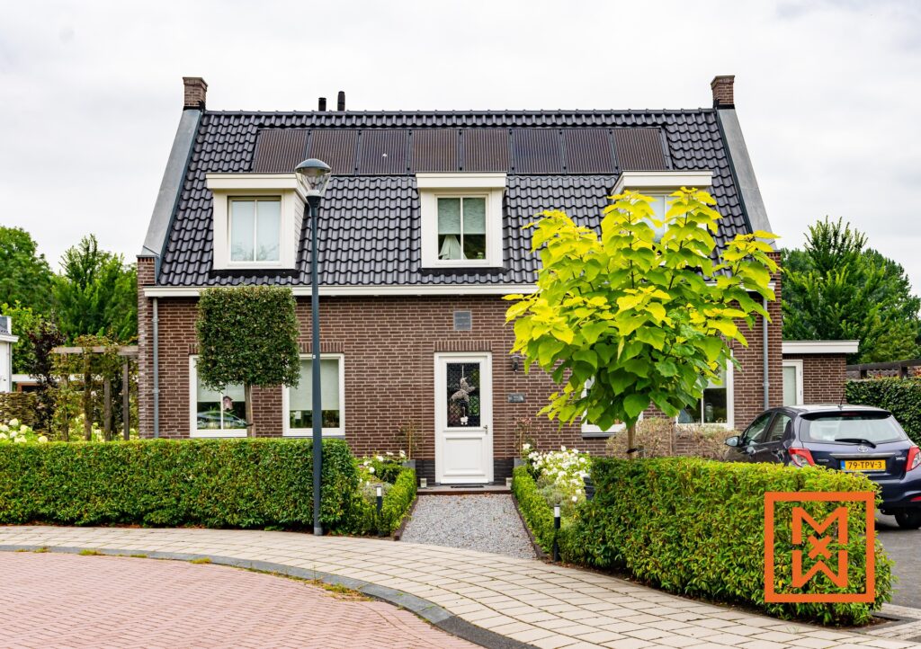 Charmant bakstenen huis met een donkergrijs pannendak en zonnepanelen, gelegen in een verzorgde tuin. Het huis heeft een witte voordeur en goed onderhouden heggen. Er staat een donkergekleurde auto in de buurt.