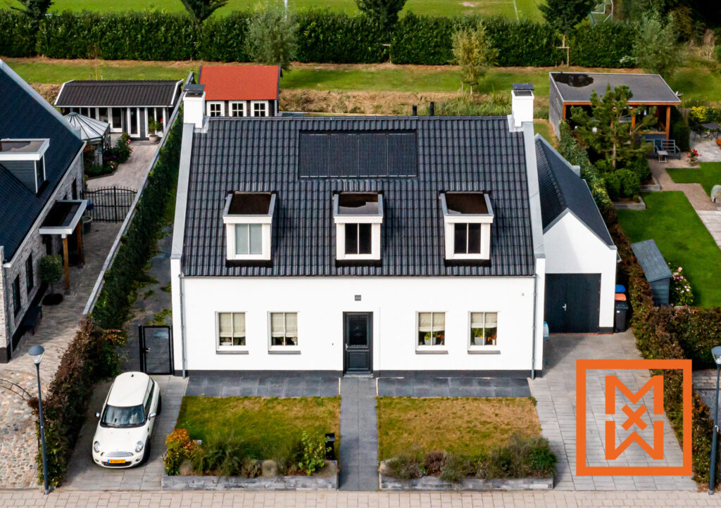 Luchtfoto van een charmant wit Nederlands huis met een donkergrijs pannendak en zonnepanelen. De woning heeft dakkapellen en een keurig aangelegde voortuin met een witte Mini Cooper ernaast. Aan de zijkant is een moderne garage zichtbaar.