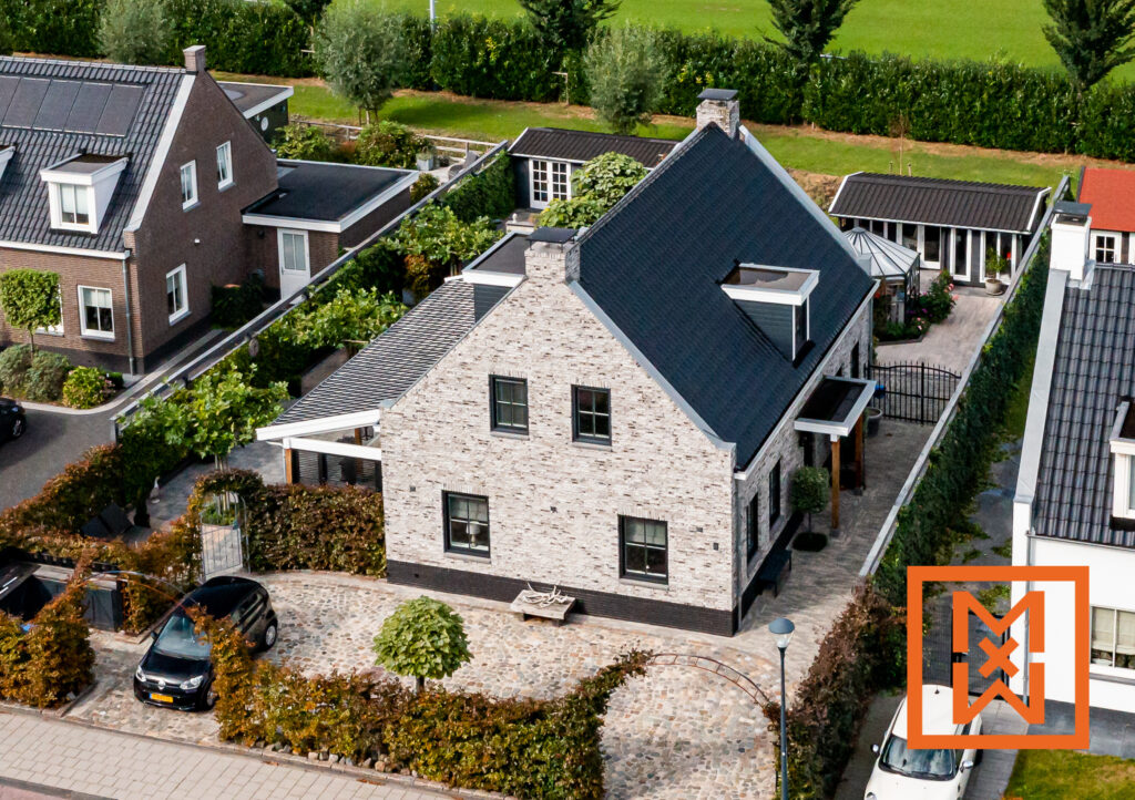 Luchtfoto van een charmant, lichtgrijs bakstenen huis met een donkergrijs dak, gelegen in een rustige woonwijk. Het huis beschikt over een zorgvuldig aangelegde tuin met hagen en een stenen oprit. Er staat een zwarte auto op de oprit.