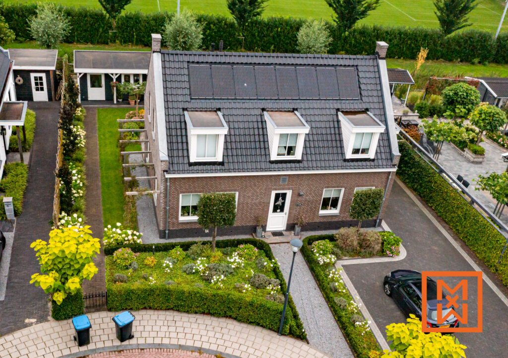 Luchtfoto van een charmant bakstenen huis met zonnepanelen op het dak, gelegen in een zorgvuldig aangelegde tuin. Het perceel beschikt over een grindpad, weelderig groen en twee donkere schuurtjes. Een donkere auto staat geparkeerd op de oprit.