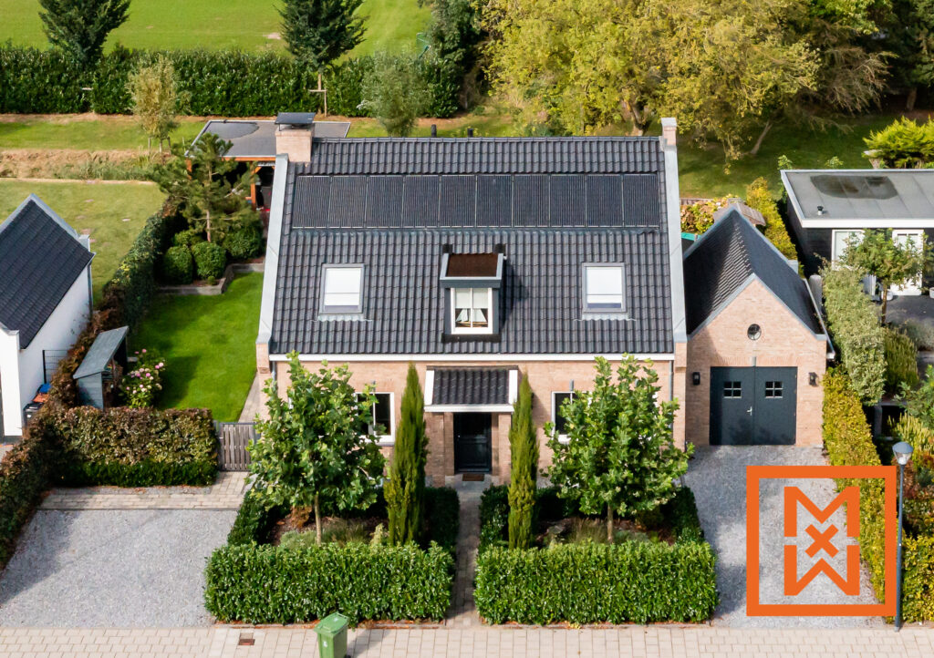 Luchtfoto van een charmant bakstenen huis met donkergrijze dakpannen en zonnepanelen, gelegen in een weelderige groene tuin. Het pand beschikt over een keurig onderhouden gazon, heggen en volwassen bomen, en een grindpad leidt naar een donkergrijze dubbele garage.