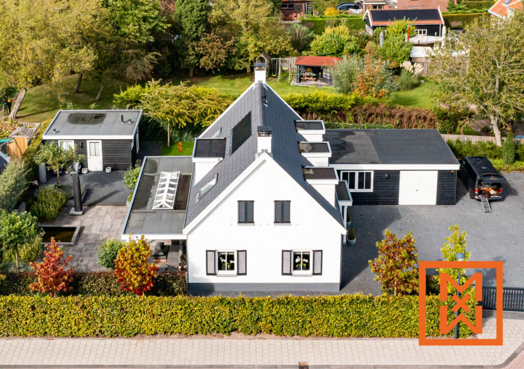 Luchtfoto van een moderne witte boerderij met zwarte accenten, omgeven door weelderige beplanting en een keurig geplaveide oprit. Het perceel beschikt over een vrijstaande garage en een kleiner bijgebouw. Herfstkleuren zijn zichtbaar in het omringende gebladerte.
