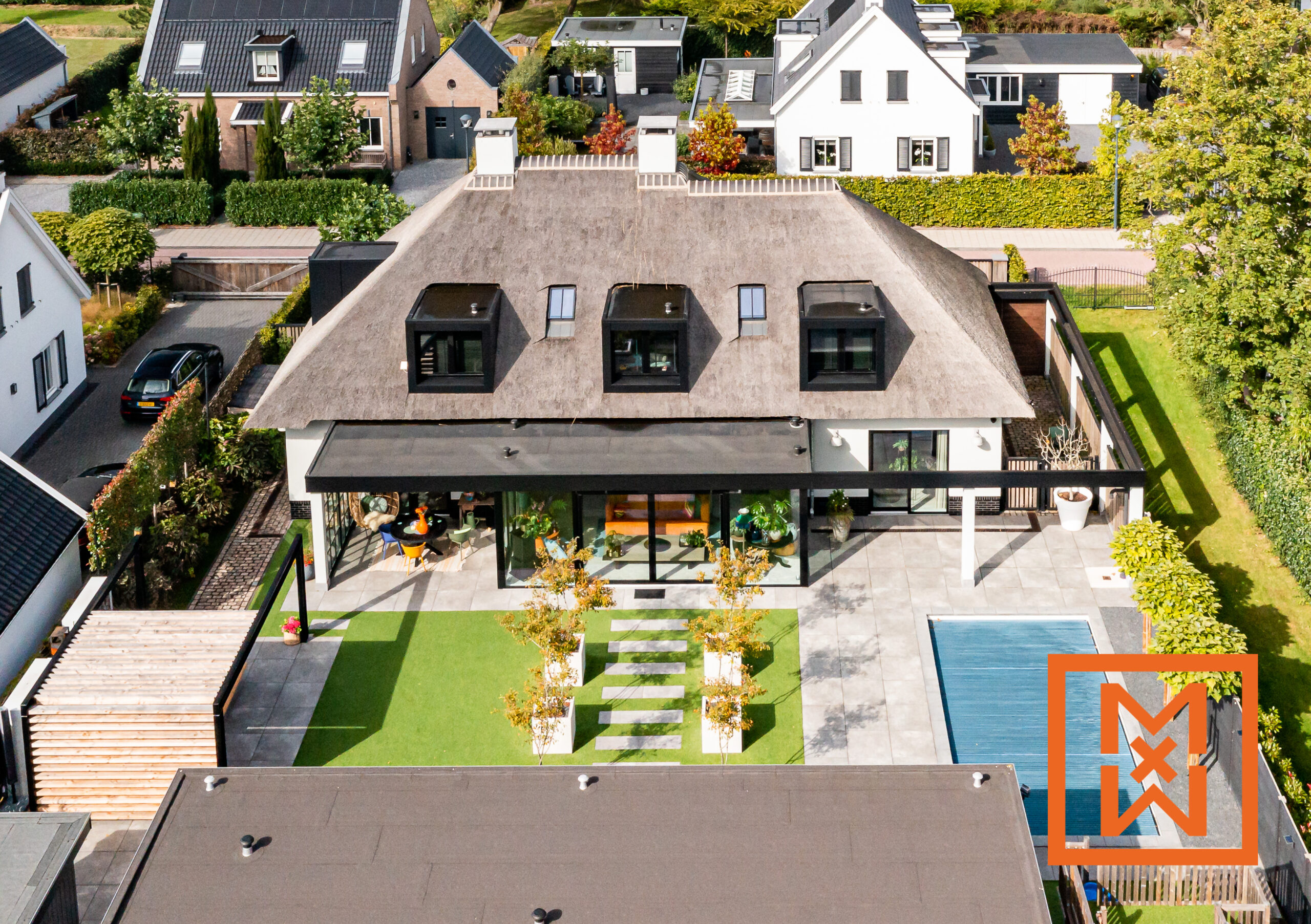 Luchtfoto van een moderne woning met rieten dak en een uitbouw met glazen wanden aan de achterzijde, met een luxueuze achtertuin met een zwembad, een verzorgd gazon en stijlvolle tuin. De woning heeft een eigentijds ontwerp dat traditionele en moderne elementen combineert.