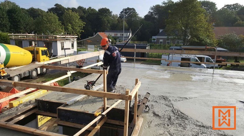Een bouwvakker gebruikt een slede om nat beton glad te maken tijdens de bouw van een nieuw huis. Het cement is lichtgrijs en de bouwvakker draagt een veiligheidshelm en een oranje overall. Op de achtergrond is een betonmixer te zien.
