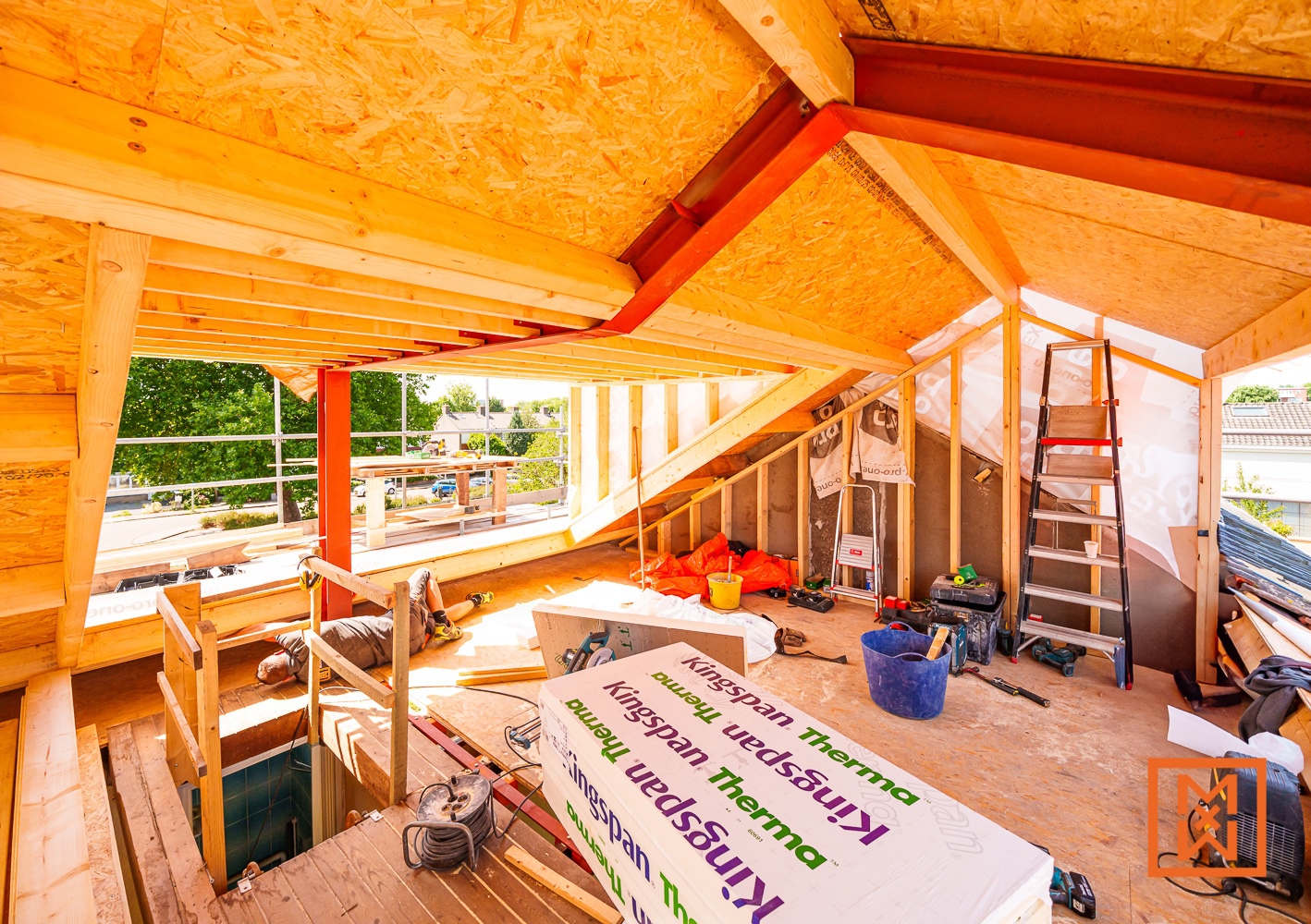Een onafgewerkte zolderkamer tijdens een dakopbouw, met zichtbare houten balken, isolatiemateriaal (Kingspan Therma) en een arbeider die pauze neemt. Gereedschap en een ladder liggen verspreid door de ruimte, wat de voortgang van de bouw benadrukt.