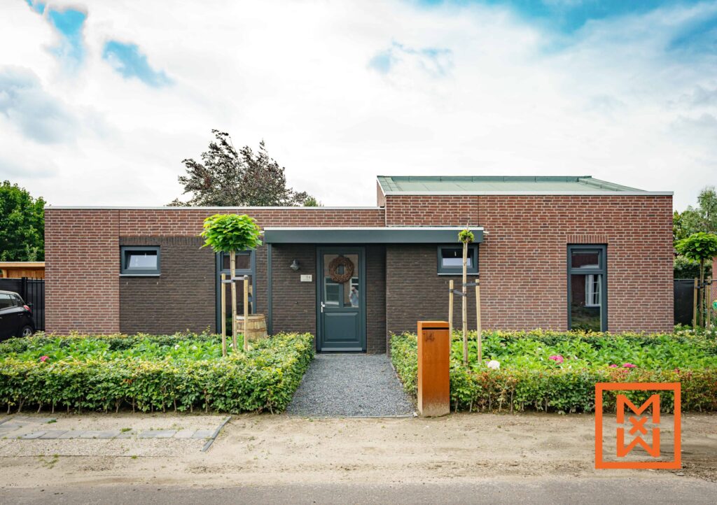 Moderne bungalow met donkergrijze voordeur en rode bakstenen gevel. Aangelegde voortuin met netjes gesnoeide heggen en jonge bomen. Brievenbus met nummer 14 zichtbaar. Rustige, voorstedelijke omgeving.