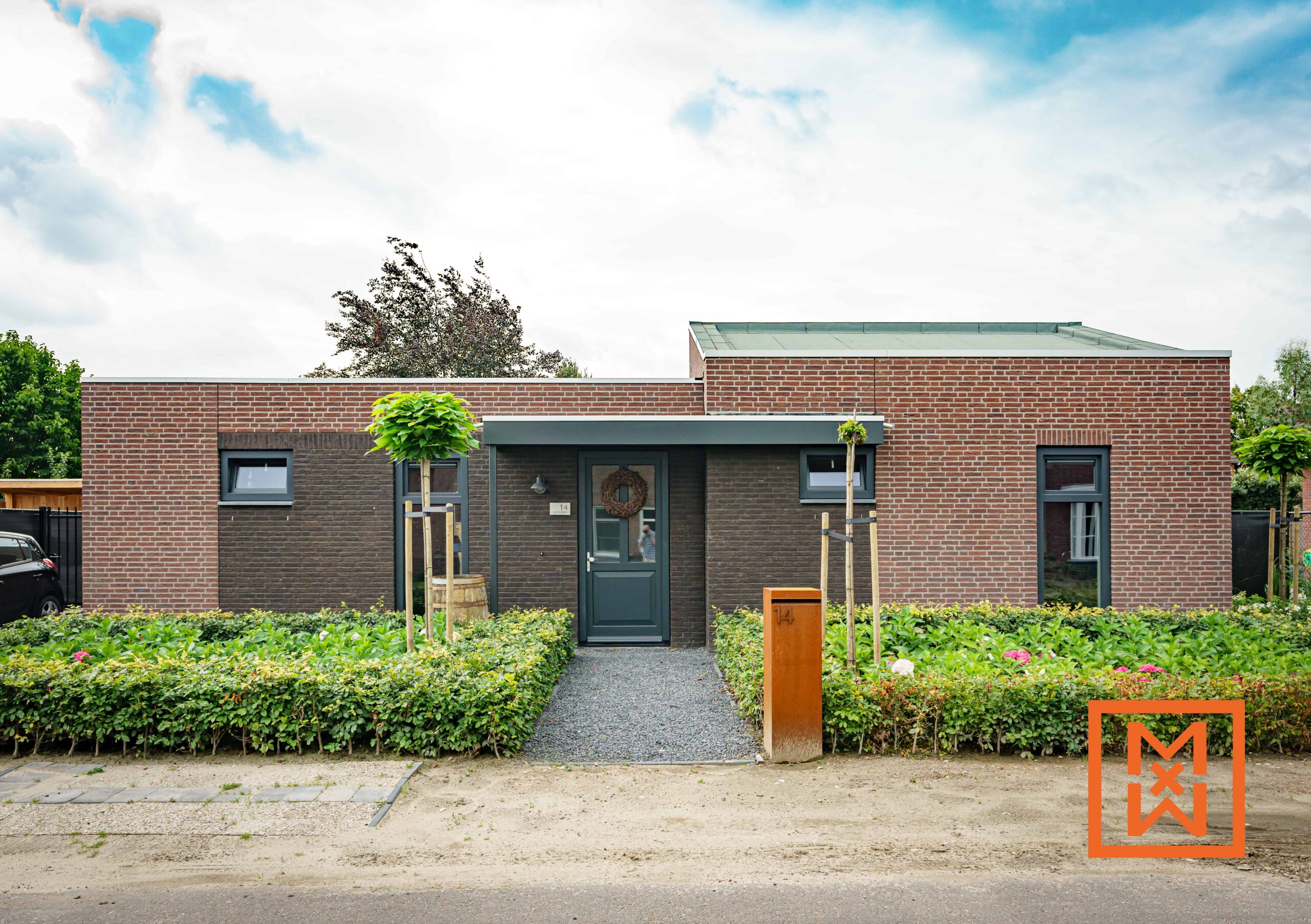 Moderne bungalow met donkergrijze voordeur en rode bakstenen gevel. Aangelegde voortuin met netjes gesnoeide heggen en jonge bomen. Brievenbus met nummer 14 zichtbaar. Rustige, voorstedelijke omgeving.