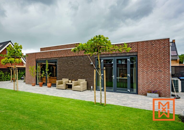 Moderne bakstenen woning met een terras en een weelderig groen gazon. Het huis heeft grote ramen en deuren met grijze kozijnen, die uitkomen op een terras met rieten meubels. Twee jonge bomen flankeren het terras. De lucht is bewolkt.