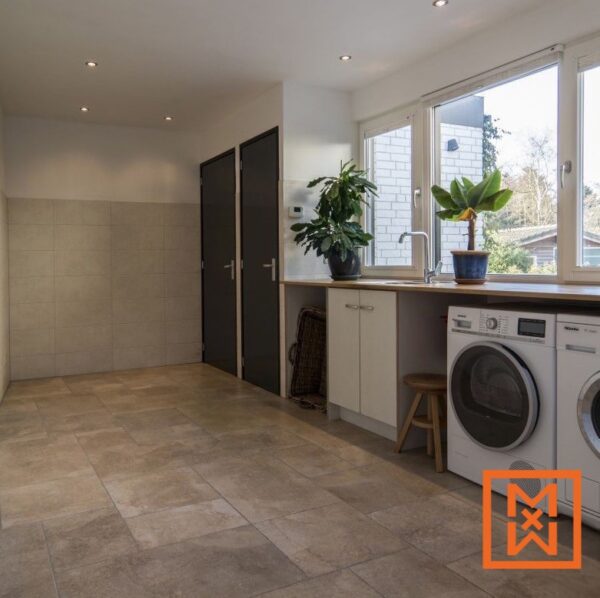 Moderne wasruimte met beige tegelvloer, witte kasten en een houten aanrechtblad. Een wasmachine en droger staan netjes naast het aanrecht, met potplanten die een vleugje natuur toevoegen. Op de achtergrond zijn twee donkere deuren zichtbaar.