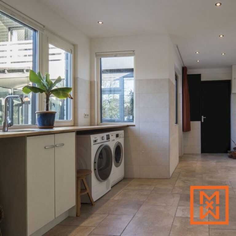 Lichte, moderne wasruimte met witte kasten, een houten aanrechtblad en een wasmachine en droger tegen een beige tegelvloer. Grote ramen zorgen voor natuurlijk licht en een bananenplant in pot zorgt voor een vleugje groen.