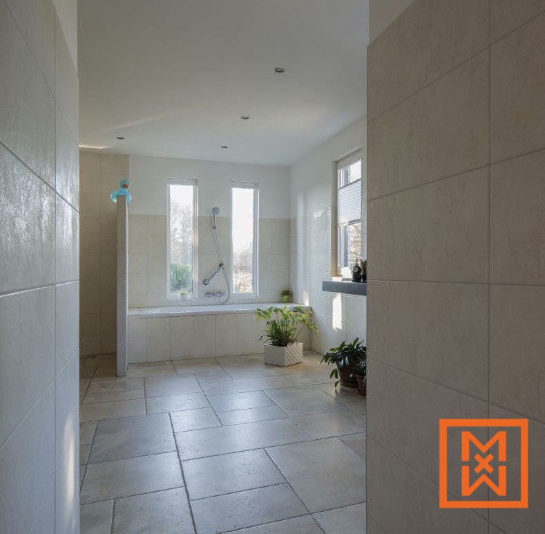 Ruime, lichte badkamer met grote beige tegels, een ingebouwd bad en douche, twee ramen en potplanten. Het rustgevende, moderne design creëert een ontspannen sfeer.