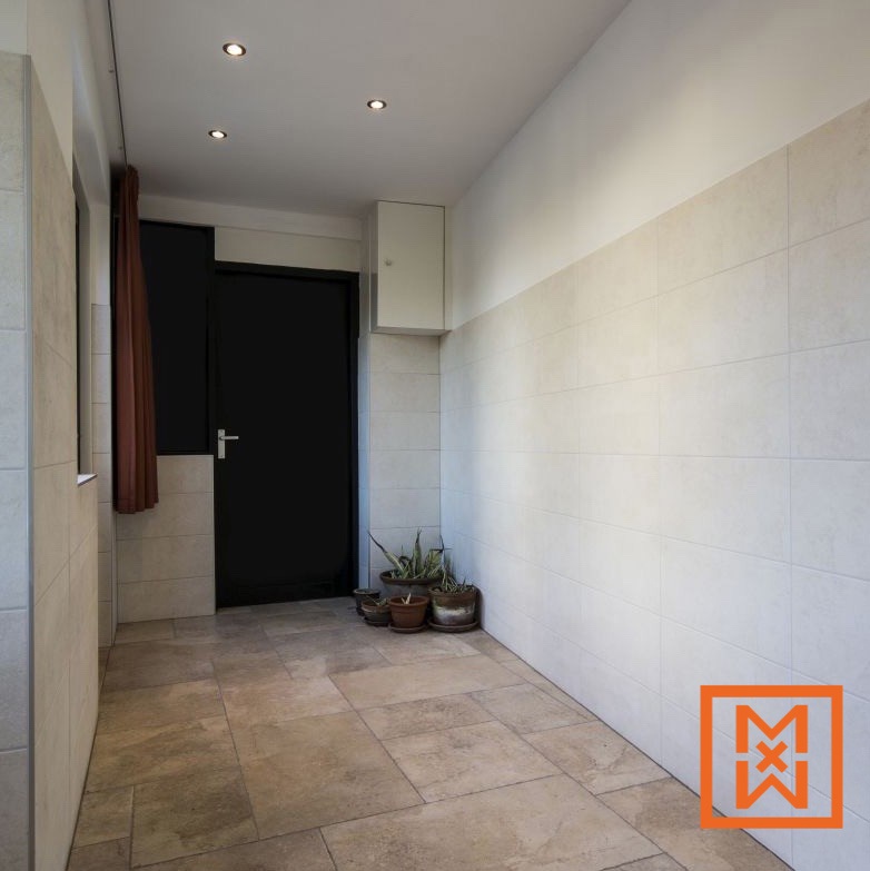 Een zonnige betegelde entree met een zwarte deur, terracotta vloertegels en potplanten. De muren zijn lichtbeige, wat een rustige en uitnodigende ruimte creëert.