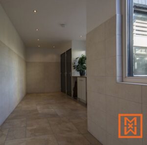 Moderne hal met beige tegelvloer en -wanden, inbouwspots en donkergrijze deuren. Een kleine plant staat op een witte kast op de achtergrond, vlak bij een raam dat natuurlijk licht binnenlaat. De algehele esthetiek is strak en minimalistisch.