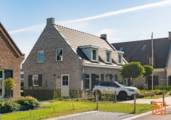 Charmante nieuwbouwwoning met een grijze bakstenen gevel, donkere luiken en dakkapellen. Een witte SUV staat geparkeerd op de oprit en de woning is omringd door weelderige beplanting. Het zonnige weer en het keurig onderhouden gazon zorgen voor een gastvrije sfeer.