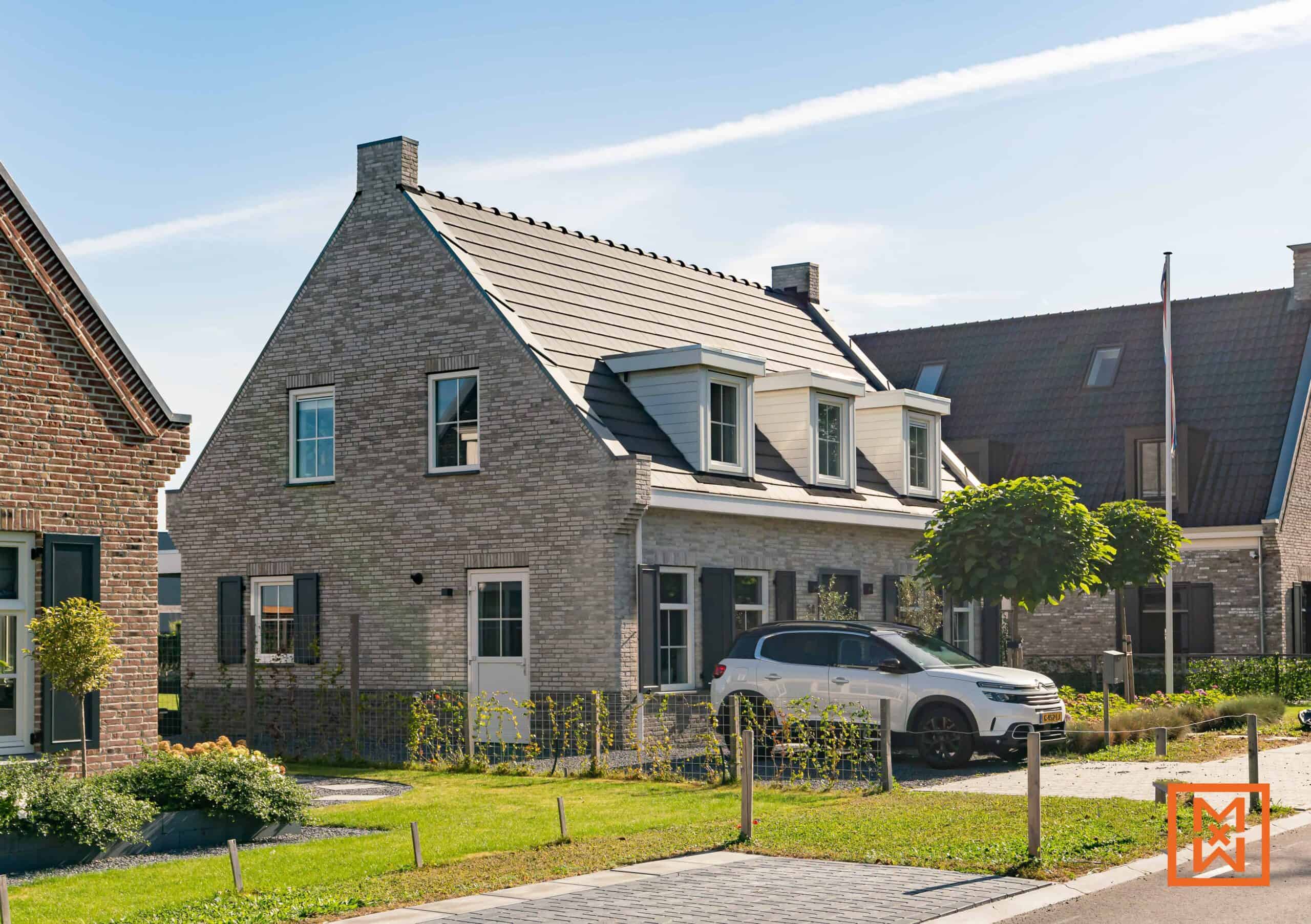Charmante nieuwbouwwoning met een grijze bakstenen gevel, donkere luiken en dakkapellen. Een witte SUV staat geparkeerd op de oprit en de woning is omringd door weelderige beplanting. Het zonnige weer en het keurig onderhouden gazon zorgen voor een gastvrije sfeer.