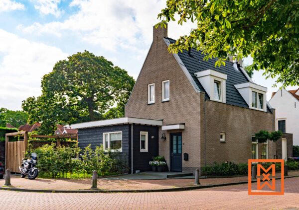 Charmant huis in Hollandse stijl met een donkerbruine bakstenen gevel en een moderne uitbouw van zwart hout. Het huis beschikt over een keurig aangelegde voortuin met rozenstruiken en een verhard pad. Er staat een motor geparkeerd in de buurt.