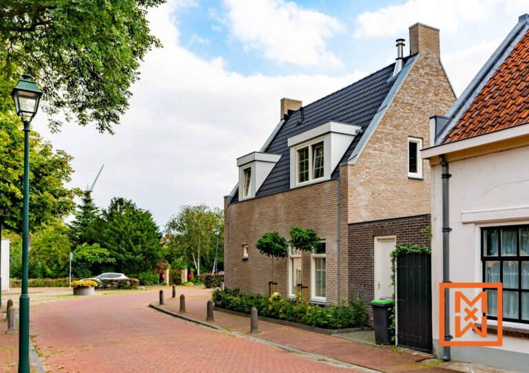 Charmante bakstenen huizen staan aan een rustige straat met klinkers, omgeven door weelderig groen en een modern huis met een donkergrijs dak. Op de achtergrond is een glimp van een windmolen te zien, wat bijdraagt aan het idyllische Hollandse dorpsgezicht.