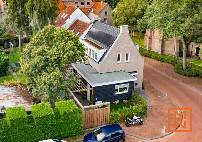 Luchtfoto van een charmant bakstenen huis met zonnepanelen, gelegen in een schilderachtig Nederlands dorpje. Het huis beschikt over een moderne uitbouw en een weelderige tuin met pergola. Een blauwe auto en motor staan vlakbij geparkeerd.