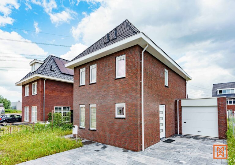 Moderne rode bakstenen woning met witte afwerking en een aangebouwde garage. De woning heeft een zadeldak en een keurig aangelegde voortuin. Een buurhuis met zonnepanelen is gedeeltelijk zichtbaar.