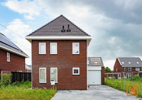 Nieuwbouw van rode baksteen met witte afwerking, een oprit met grijze bestrating en een witte garagedeur. Het huis heeft zonnepanelen op het dak en een keurig onderhouden gazon. De lucht is gedeeltelijk bewolkt.
