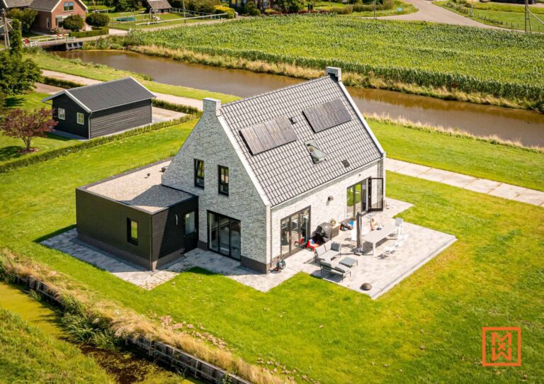 Luchtfoto van een modern, lichtgrijs bakstenen huis met zwarte accenten en zonnepanelen op het dak, gelegen in een groen landschap naast een kanaal. Het huis beschikt over een ruim terras met tuinmeubilair.