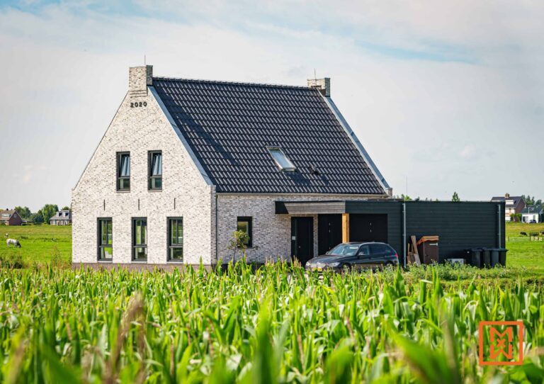 Moderne, witte bakstenen boerderij met donkergrijs dak, gelegen in een groen korenveld. Een zwarte auto staat geparkeerd op de oprit en het jaartal "2020" is zichtbaar op het metselwerk. De woning heeft grote ramen en een eigentijds design.