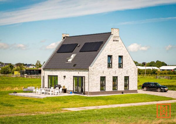 Moderne boerderij met zwarte dakpannen en zonnepanelen, met een lichtgrijze bakstenen buitenkant. Het huis heeft grote ramen met zwarte kozijnen en een terras met tuinmeubilair. Een zwarte auto staat geparkeerd op de oprit, tegen een weelderig groen veld. "2020" staat subtiel op de gevel.