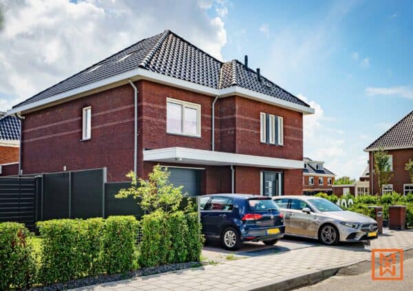 Moderne, twee verdiepingen tellende bakstenen woning met een donkergrijs dak en auto's geparkeerd op de oprit. Het huis heeft een strak ontwerp, een kleine voortuin met een heg en een donkergrijze schutting. Een zonnige dag met een blauwe lucht en zachte wolken.