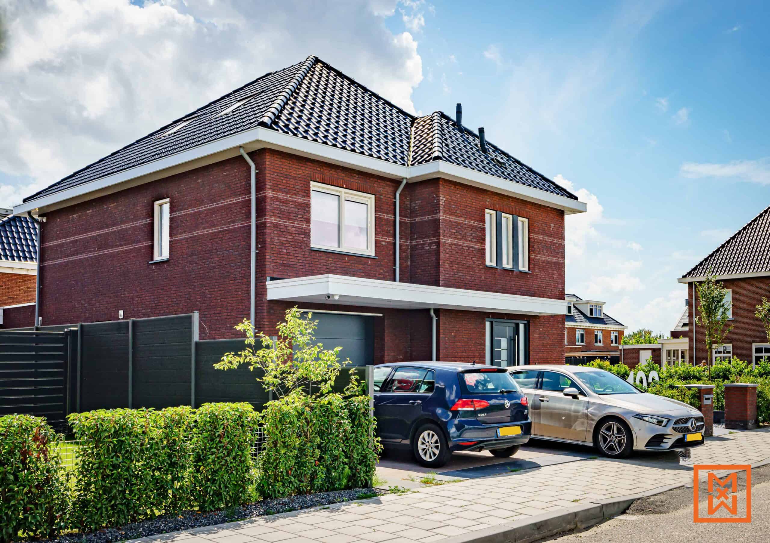 Moderne, twee verdiepingen tellende bakstenen woning met een donkergrijs dak en auto's geparkeerd op de oprit. Het huis heeft een strak ontwerp, een kleine voortuin met een heg en een donkergrijze schutting. Een zonnige dag met een blauwe lucht en zachte wolken.
