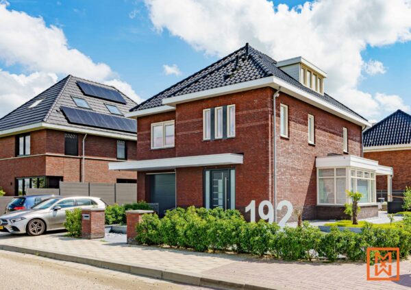 Moderne bakstenen woning met prominent aanwezige zonnepanelen op buurhuis 192. Aangelegde voortuin met netjes gesnoeide heggen. Een zilveren auto staat geparkeerd op straat.