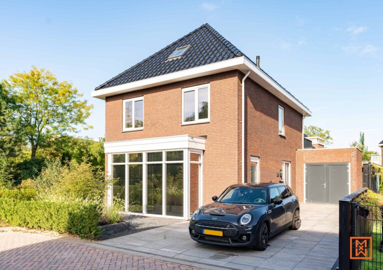 Moderne bakstenen woning met twee verdiepingen en een donkergrijze Mini Cooper op de oprit. De woning beschikt over een serre en een vrijstaande garage. Weelderig groen omringt de woning onder een strakblauwe hemel.