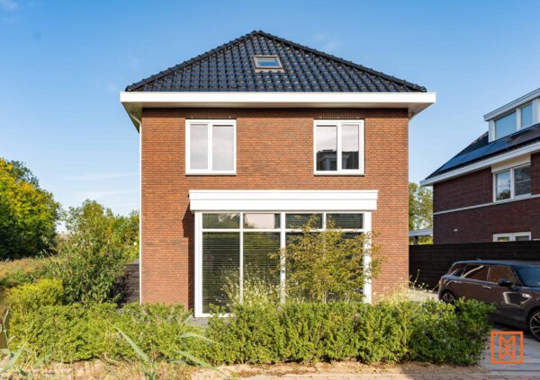 Modern bakstenen huis met een zwart dak en grote ramen, een toonbeeld van eigentijds woningontwerp. De aangelegde tuin met weelderig groen omringt het huis en creëert een rustige sfeer.