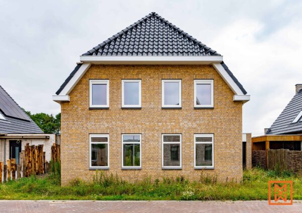 Nieuwbouwwoning van gele baksteen met een donkergrijs dak en witte kozijnen. Het huis heeft meerdere ramen op gelijke afstand van elkaar. De voortuin is onverhard en heeft een onregelmatige grasmat.