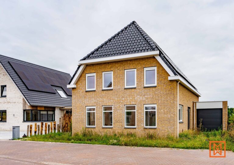 Nieuw gebouwd, lichtgeel bakstenen huis met een donkergrijs pannendak, moderne ramen en een strakke aangebouwde garage. Het huis staat aan een geplaveide straat, met hoog gras op de voorgrond en een buurhuis met gedeeltelijk zichtbare zonnepanelen.